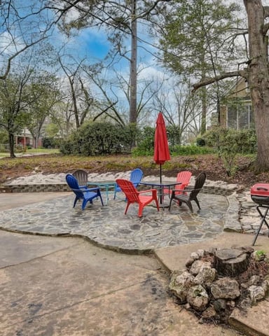 Backyard patio with colorful Adirondack chairs, red umbrella, grill, and stone fire pit surrounded by tall trees
