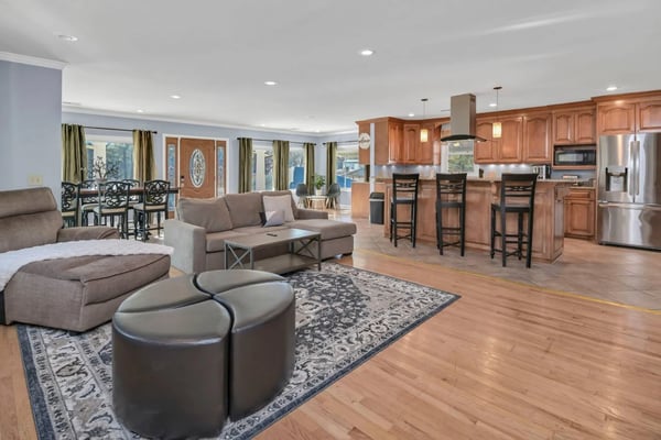 Open-plan living room and kitchen with wood flooring, beige sofa, leather ottomans, wood cabinetry, stainless steel appliances, and lake views through large