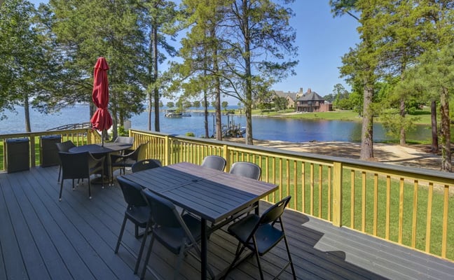 Waterfront deck with dining table, yellow railing, red umbrella, and view of lake with trees and distant house